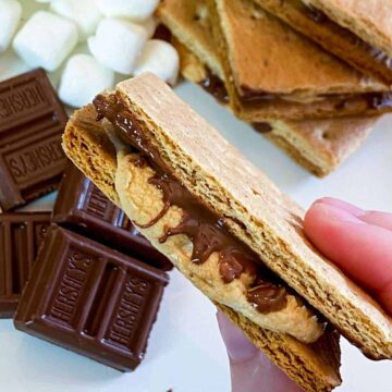 Holding one of the air fryer s'mores over chocolate, marshmallows, and a stack of s'mores