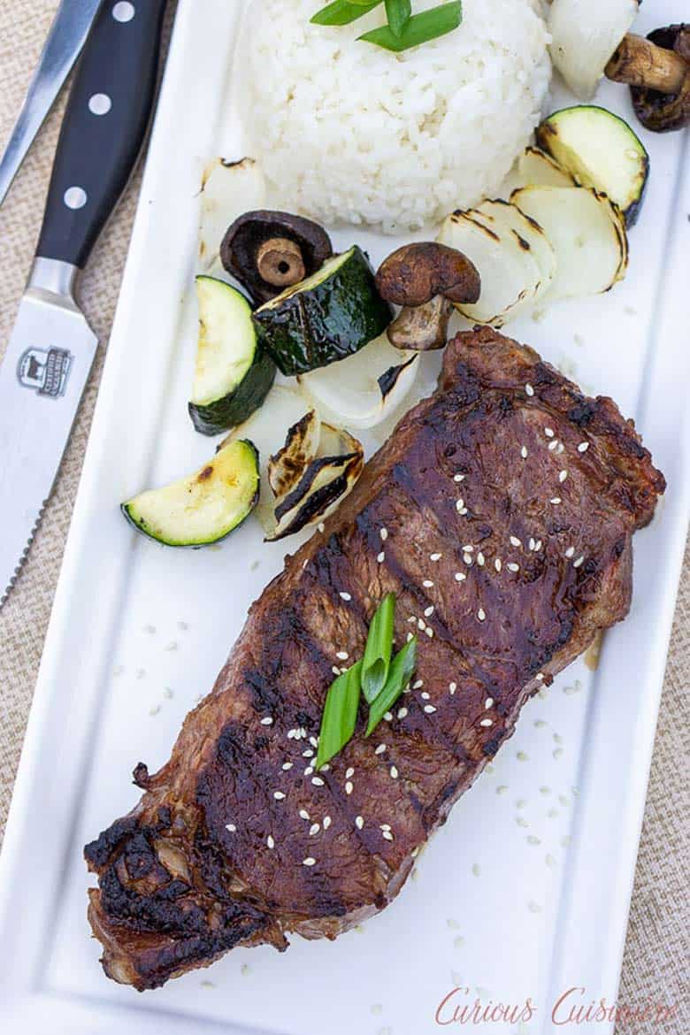 A Japanese grilled hibachi steak on a white rectangular platter with a side of white sticky rice, mushrooms, and sliced grilled zucchini