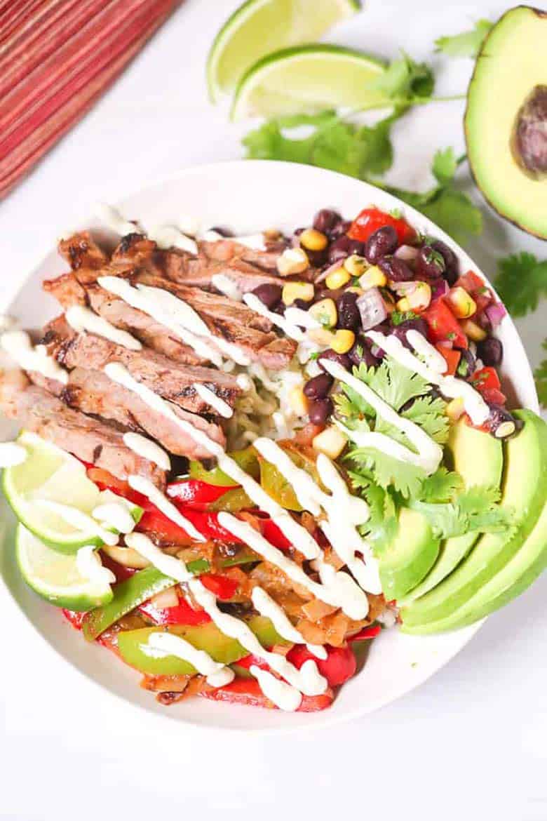 Top-down view of a grilled steak fajita bowl with avocado slices, crema, and lime wedges, next to half an avocado and some more lime wedges