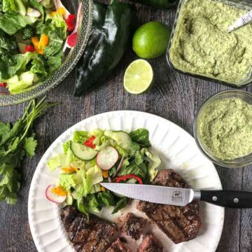 Grilled steaks with creamy poblano sauce on a white plate with a knife surrounded by guacamole, limes, and a salad on a table
