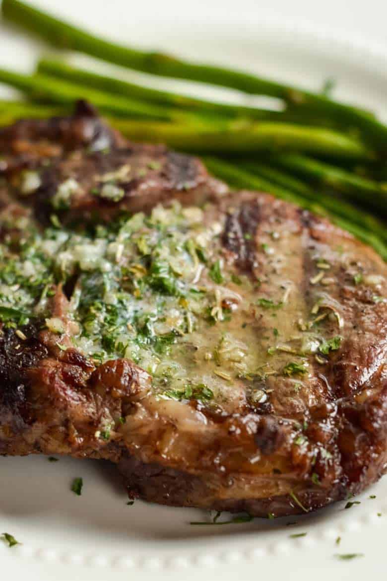 Ribeye steak on a white plate with a side of asparagus and garlic butter melted on top