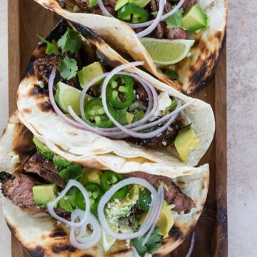 Three honey lime steak tacos topped with sliced red onions, avocado, and lime wedges on grilled tortillas resting on a cutting board