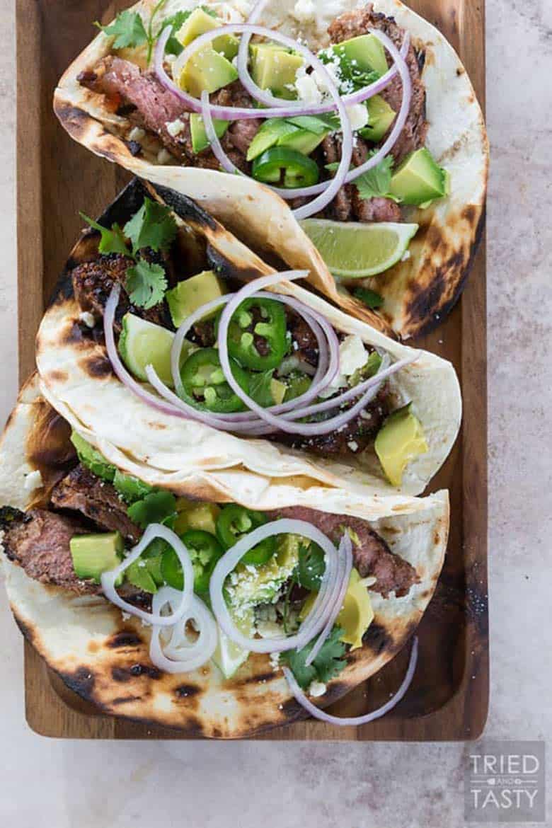 Three honey lime steak tacos topped with sliced red onions, avocado, and lime wedges in grilled tortillas resting on a cutting board