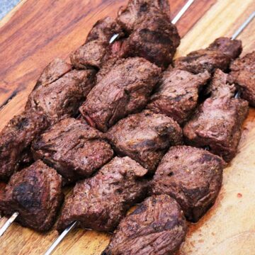 Four cumin rubbed steak kabobs on skewers on a light wooden surface