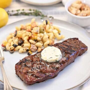 A grilled strip steak on a white plate with pasta salad on the side topped with herb compound butter