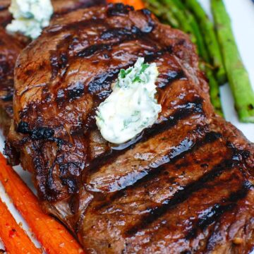Close up of two lemon garlic butter grilled ribeyes on top of each otheer on a plate with asparagus and whole cooked carrots on the side