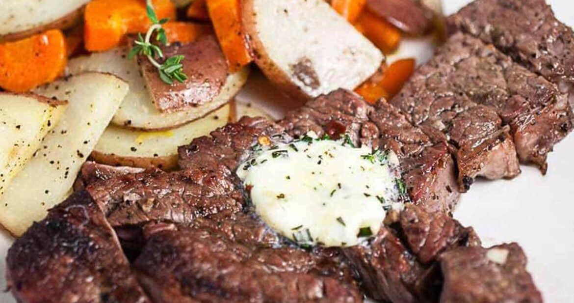 Grilled chuck steak on a white plate topped with garlic butter with a side of potatoes and carrots