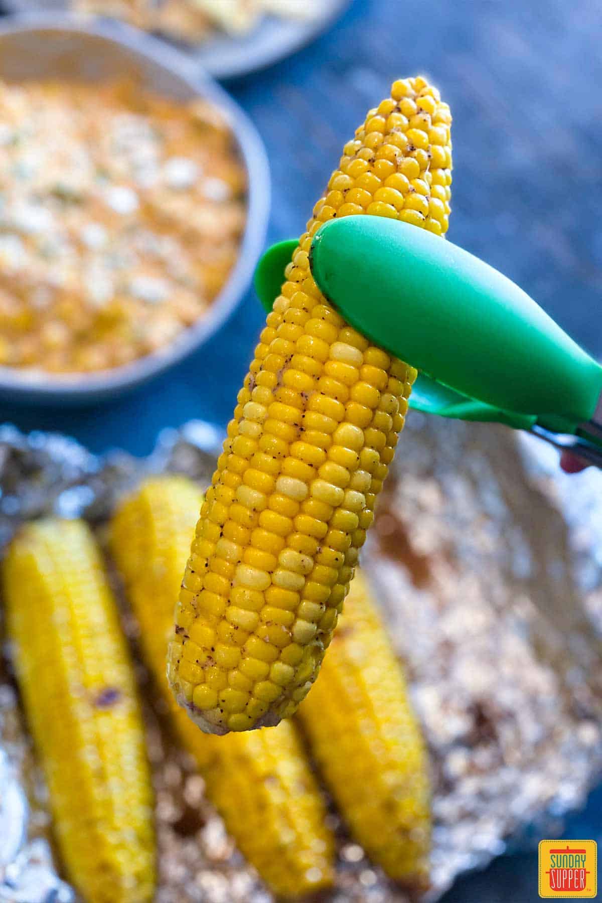 Lifting a corn cob up with silicone tongs over the foil pack of three corn cobs