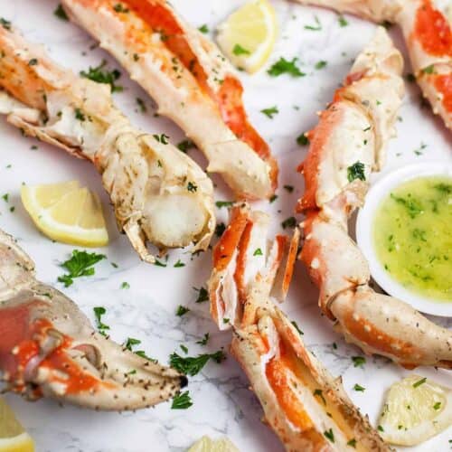 Grilled crab legs on a white surface with fresh herbs and a side of garlic butter in a little bowl