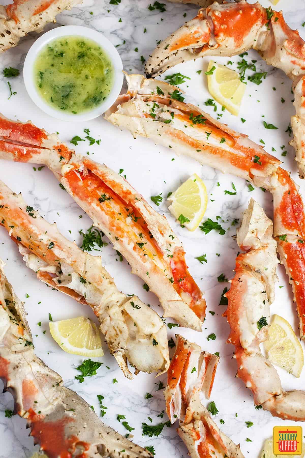 Grilled crab legs on a white surface with a cup of garlic butter sauce and fresh herbs sprinkled on top