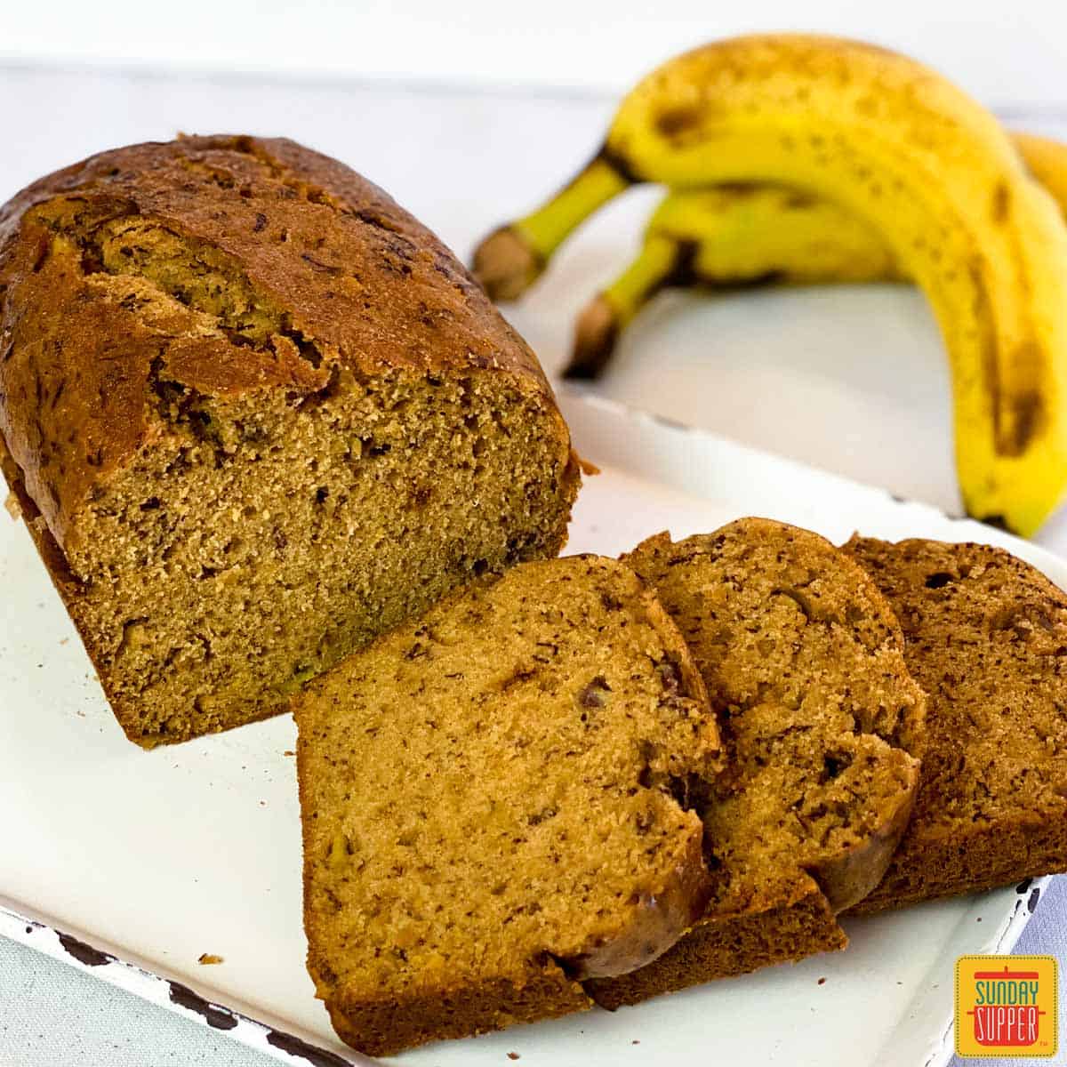 Three slices of banana bread next to a loaf of Instant Pot Banana Bread on a white platter with two bananas in the background