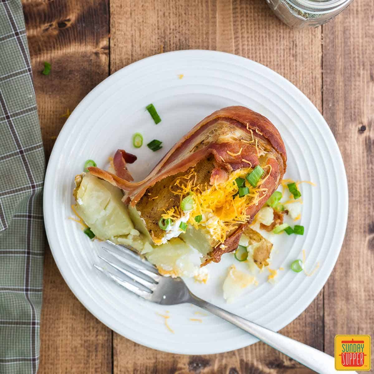 A bacon-wrapped potato on a white plate with a fork, split open and stuffed with cheese, sour cream, and green onions