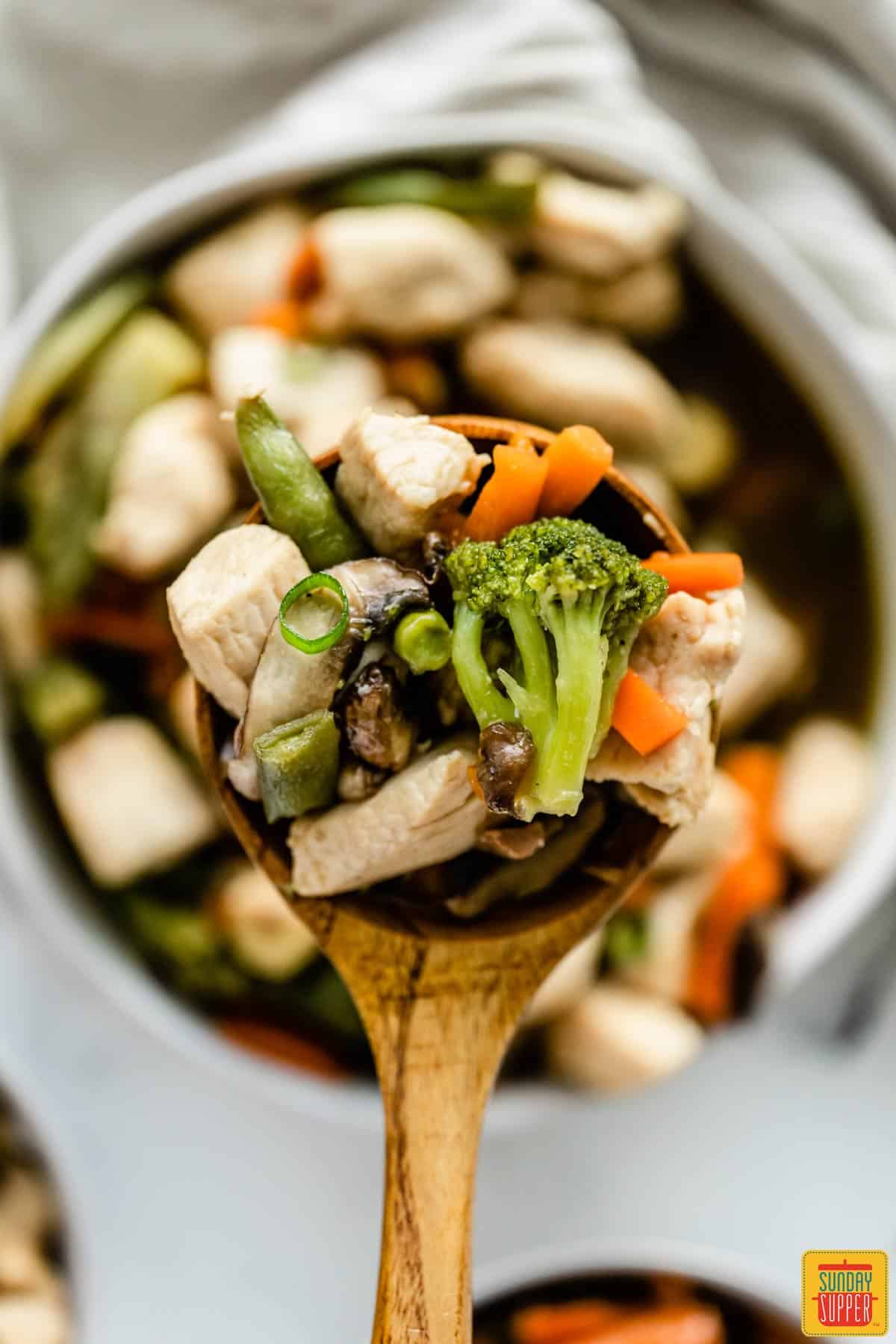 A wooden spoon with a serving of chicken stir-fry lifted out of a bowl in the background
