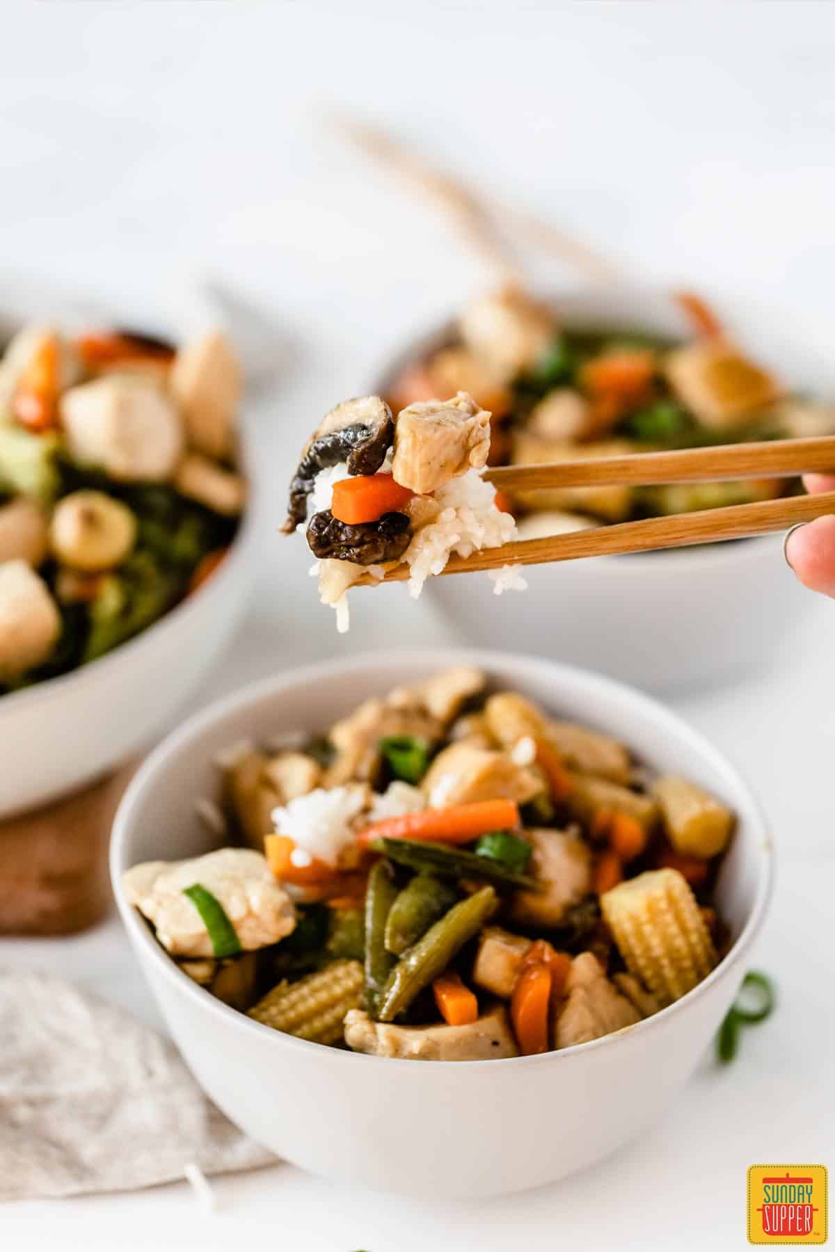 Lifting a bite of 5-ingredient chicken stir-fry out of a white bowl with a pair of chopsticks