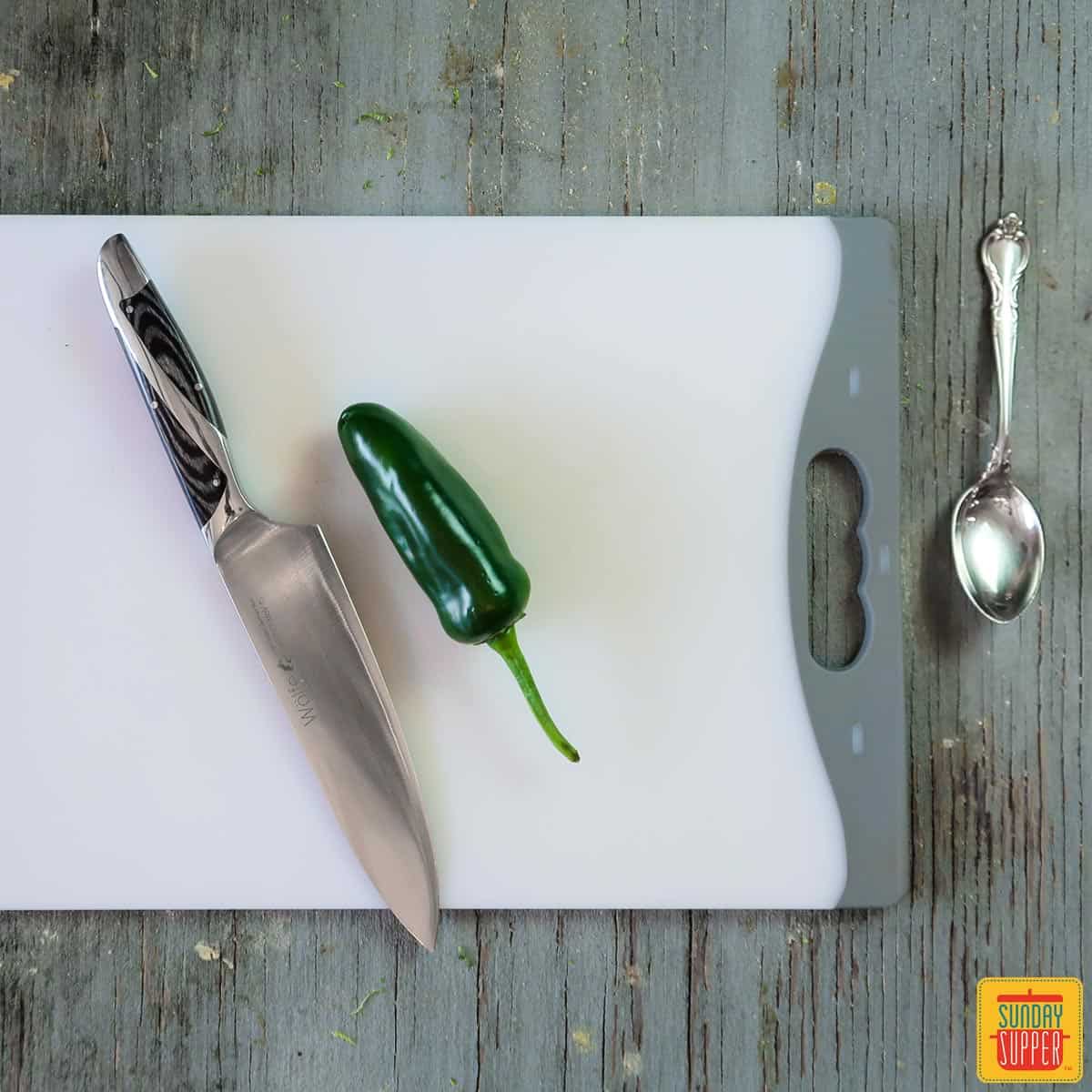 A cutting board with a jalapeno and knife next to a spoon, getting ready to show you how to cut a jalapeno