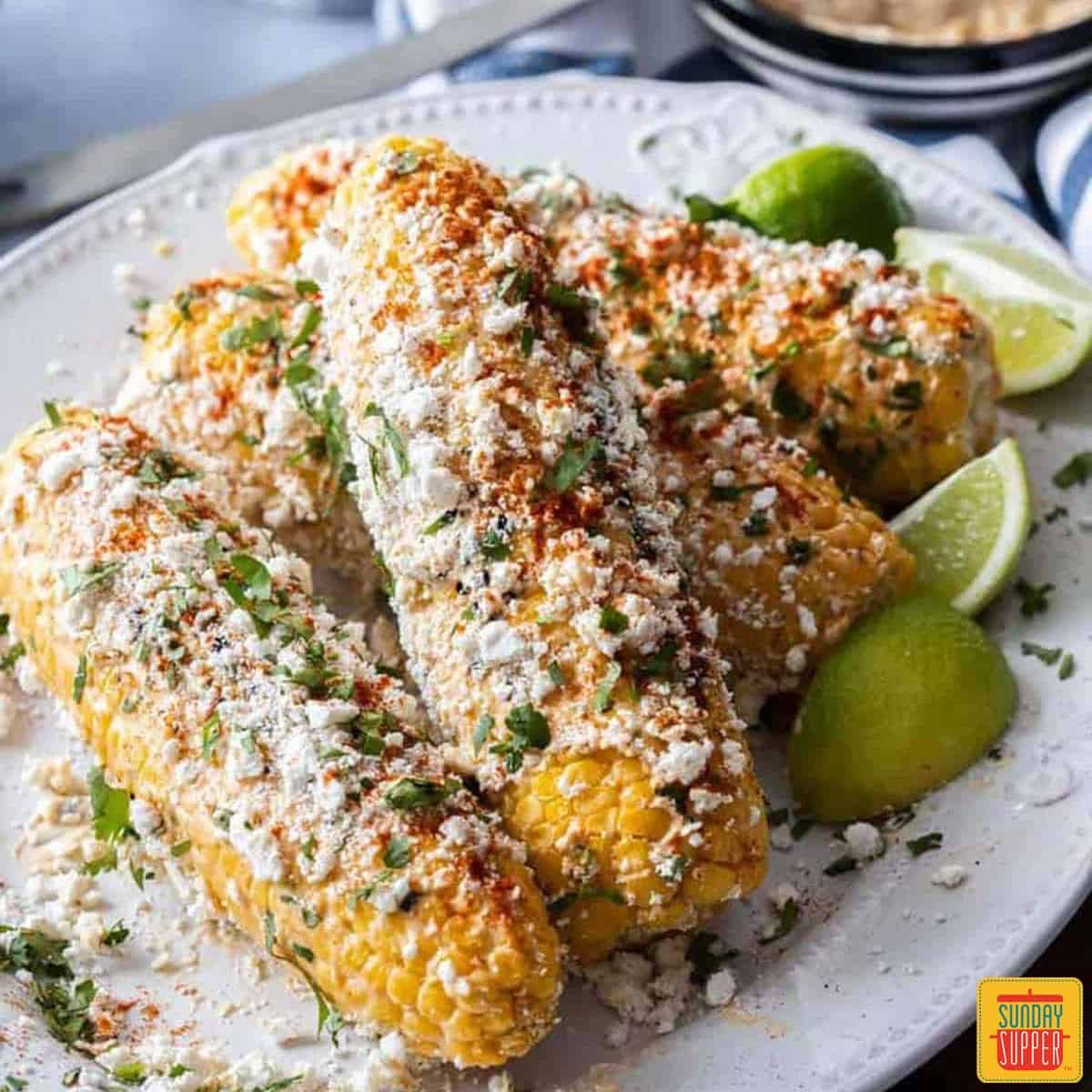 Elotes Mexicanos piled on a white platter with lime wedges