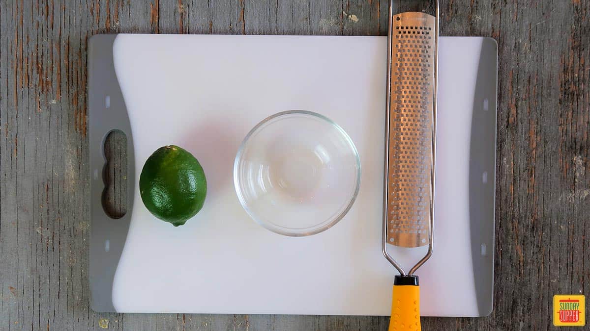 The tools you need for how to zest a lime: a grater, bowl, and lime, on a cutting board