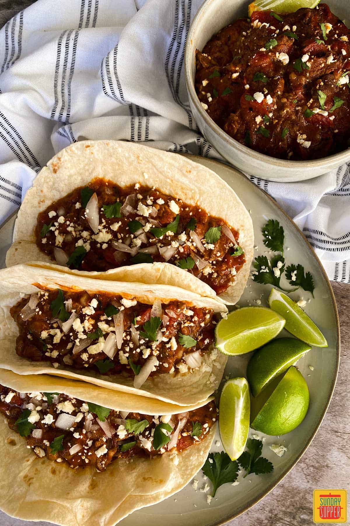 Three carne picada tacos next to a bowl of carne picada and limes