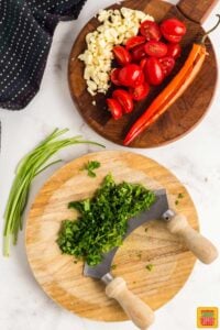 Chopped parsley next to sliced red chili pepper, minced garlic, and halved cherry tomatoes on two separate wooden boards