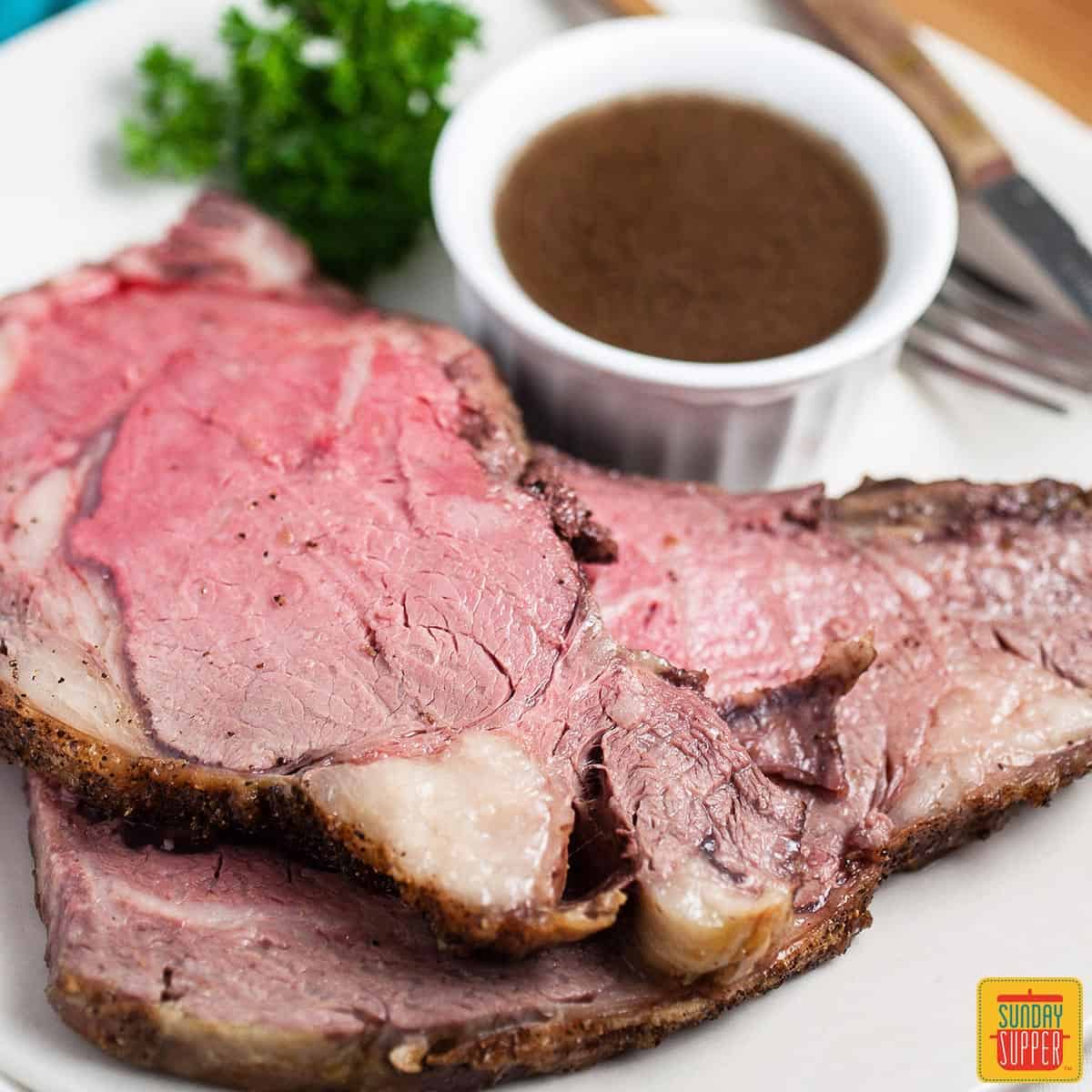 Slices of boneless prime rib on a white plate next to herbs and a ramekin of au jus