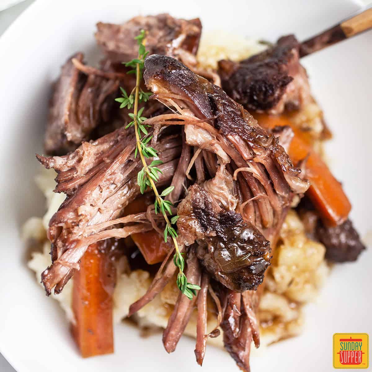 Close up of dutch oven pot roast in a white dish with a sprig of rosemary