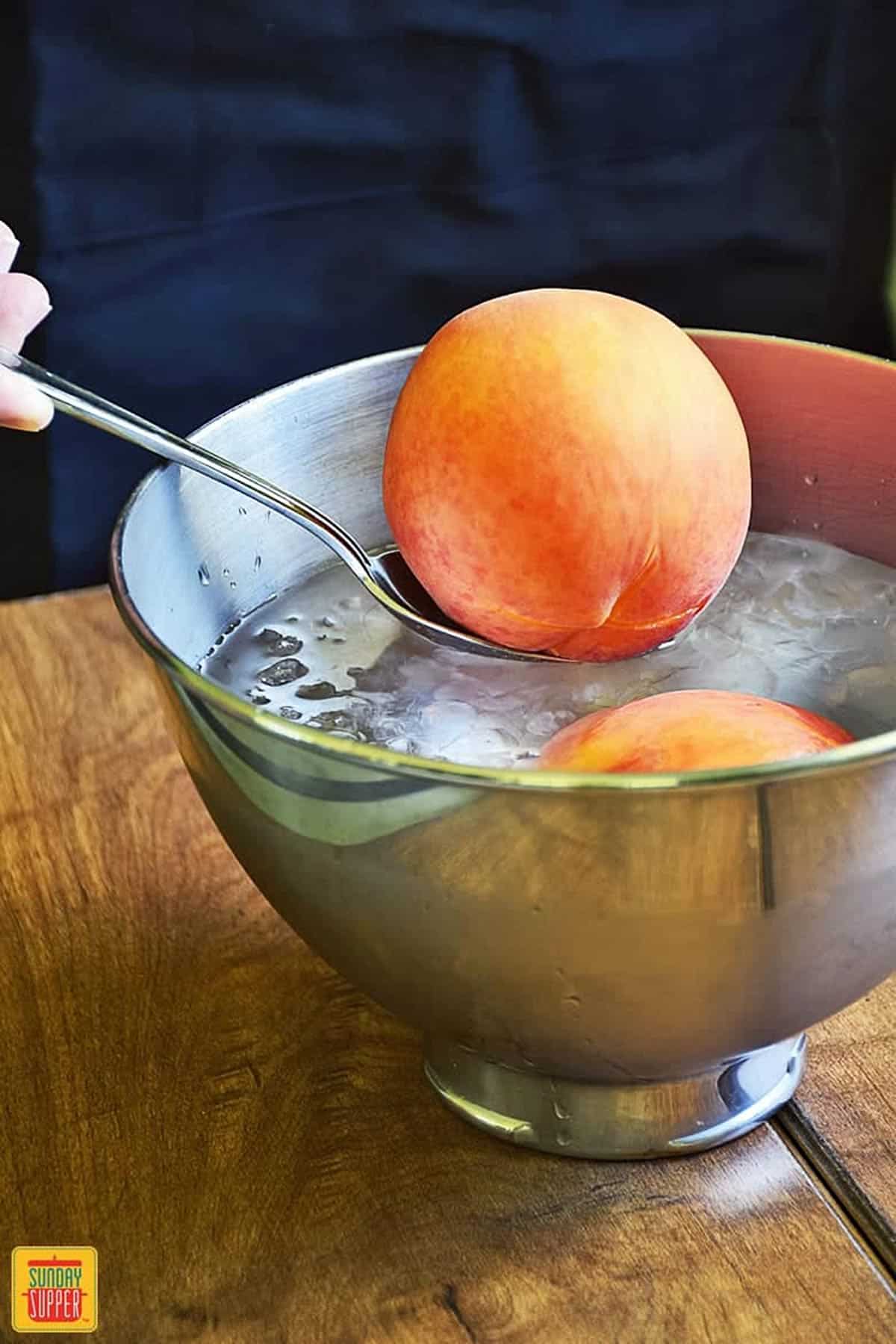 Adding a peach on a spoon to an ice water bath in a metal bowl