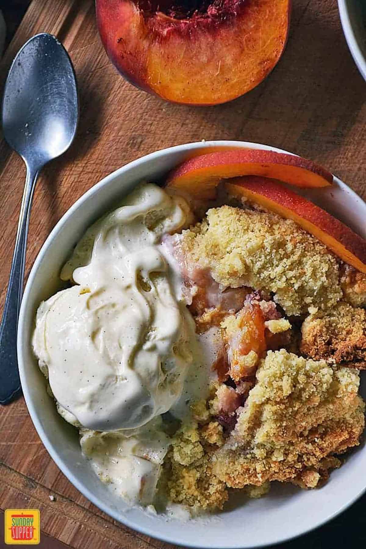 A white bowl with peach cobbler with cake mix, fresh peach slices, and ice cream next to half a peach and a spoon