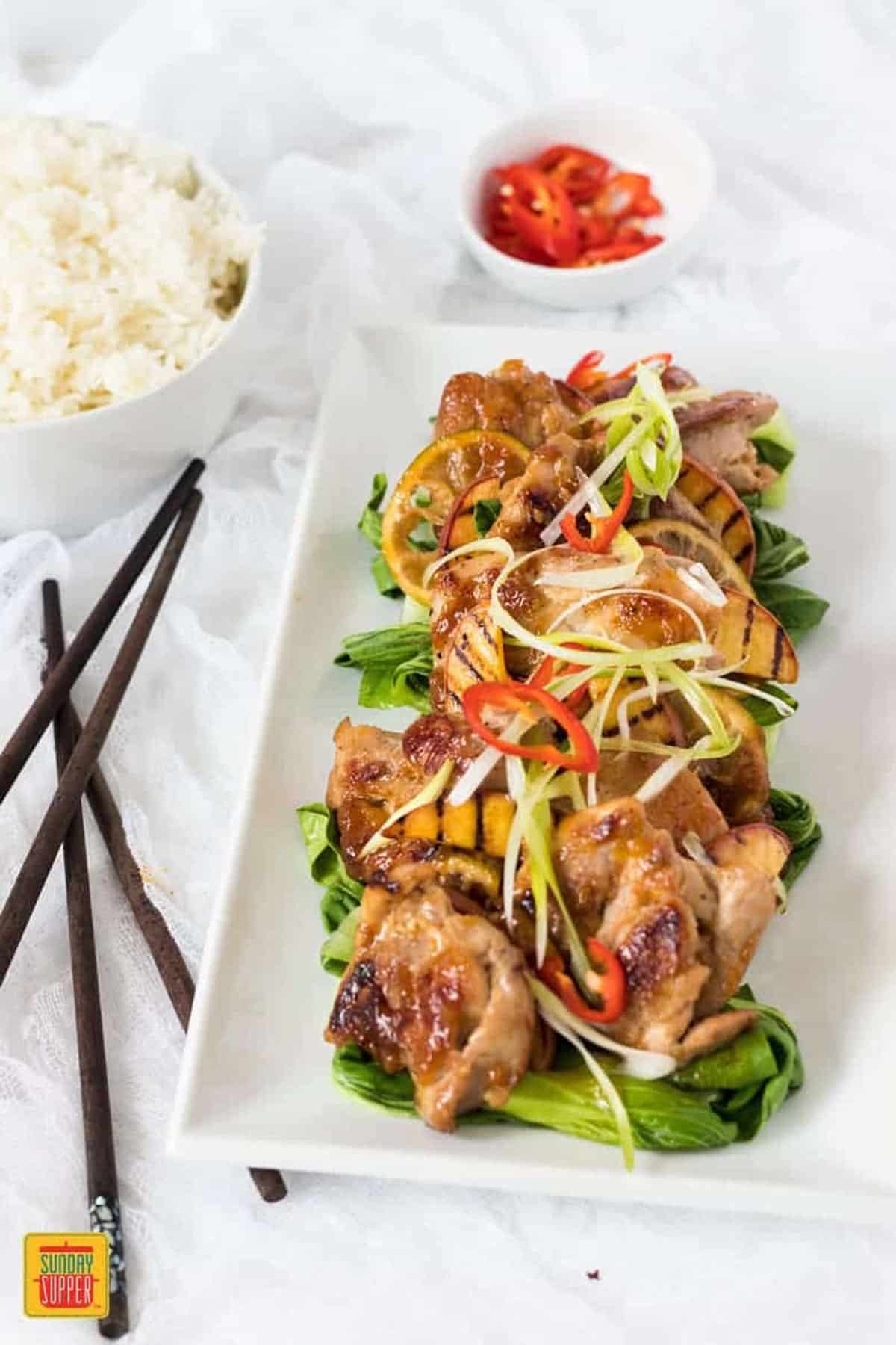 Peach glaze chicken on a white platter next to rice, peppers, and chopsticks