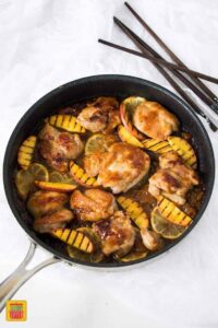 Peach glaze chicken cooking in a skillet with peaches and citrus next to chopsticks