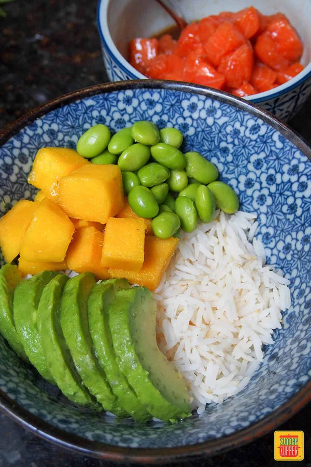 Sliced avocado, diced mango, and edamame over rice in a white and blue bowl
