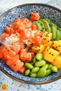 A forkful of diced salmon in a salmon poke bowl next to mango, edamame, and sliced avocado
