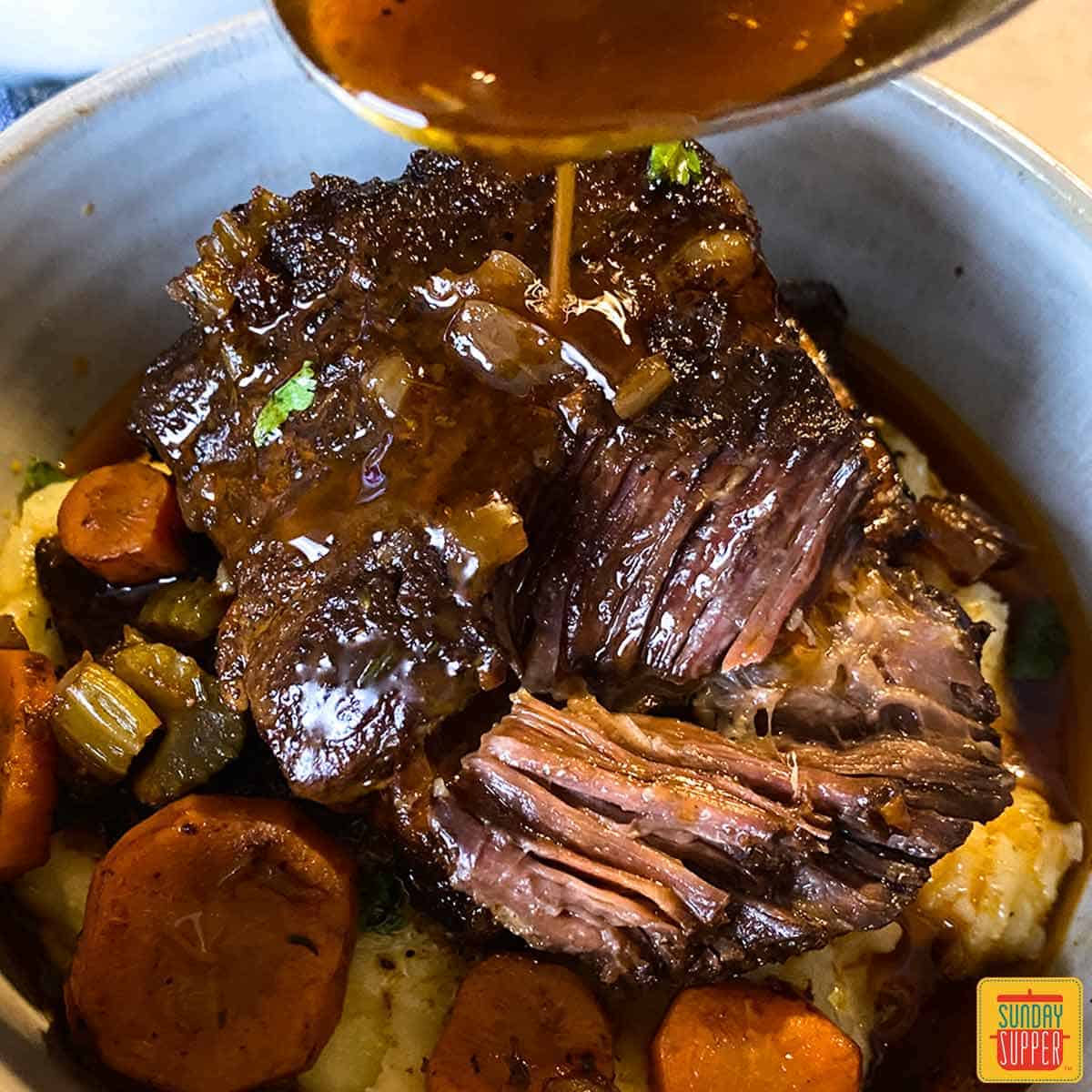 Pouring gravy onto short ribs in a bowl ov er vegetables