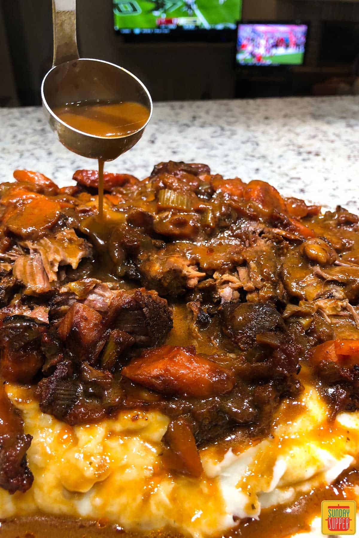 pouring sauce onto a beef chuck roast with a football game on TV in the background