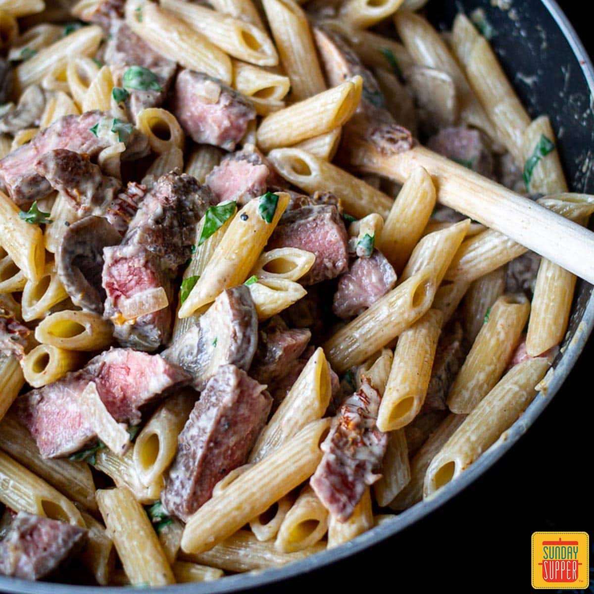 Creamy penne pasta up close in a skillet with a wooden spoon and slices of prime rib