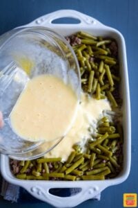 Pouring egg mixture over green beans and ground beef in casserole dish