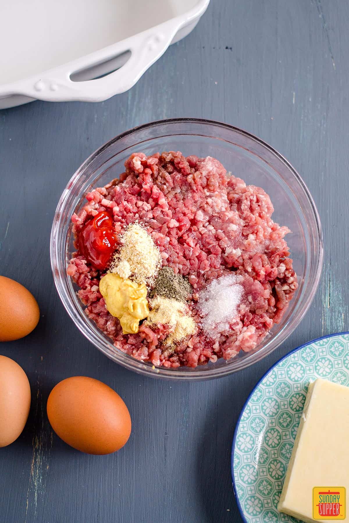 Ingredients to make keto cheeseburger casserole on blue surface and in a bowl