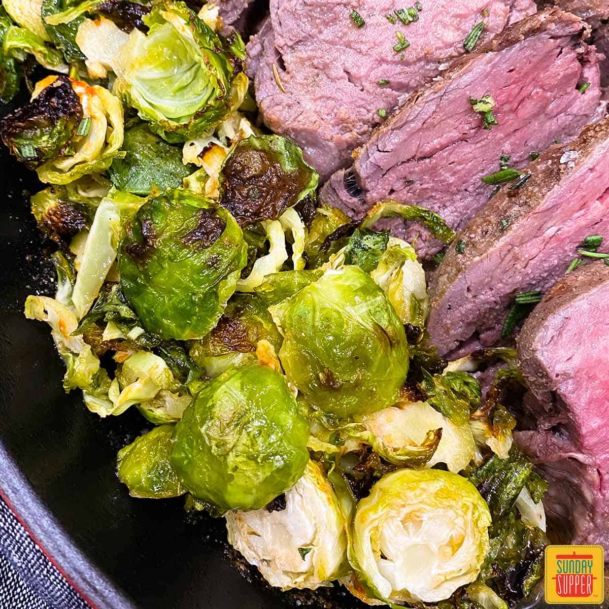 Close up of roasted brussels sprouts next to sous vide tenderloin in a skillet