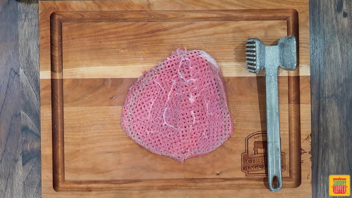 A tenderized steak on a cutting board next to the meat mallet