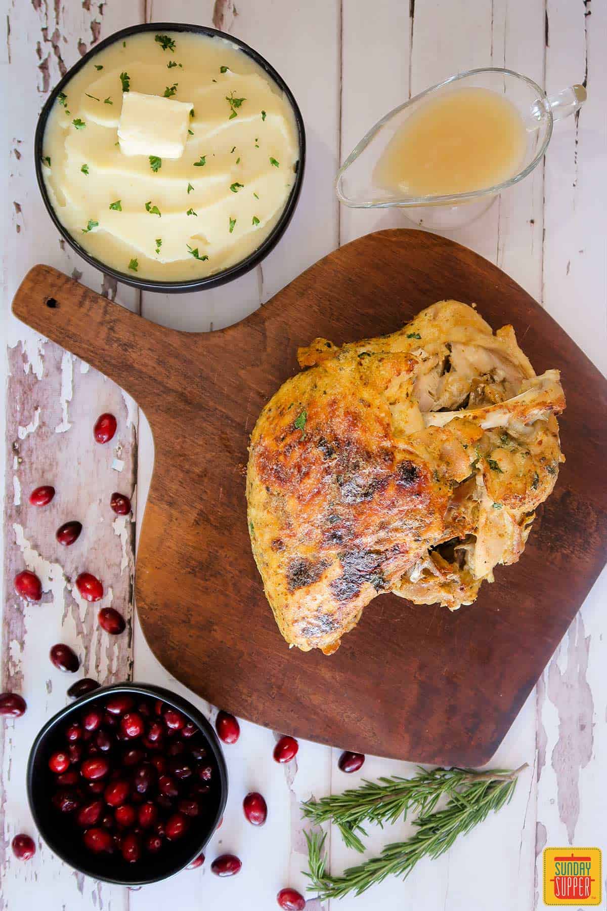 Instant pot turkey breast on a cutting board next to a bowl of cranberries, mashed potatoes, rosemary sprigs, and gravy