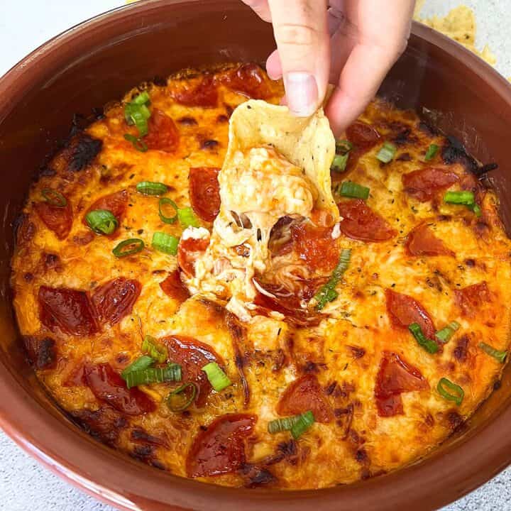 pizza dip in a brown baking dish being scooped by a chip