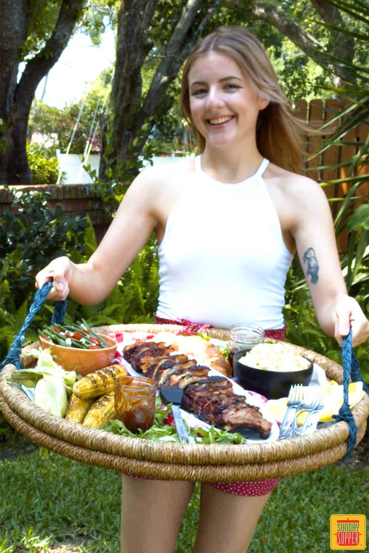 Smiling woman holding tray of baby back ribs and side dishes outside