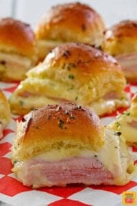 Several ham and cheese sliders ready to eat on a red and white checked paper