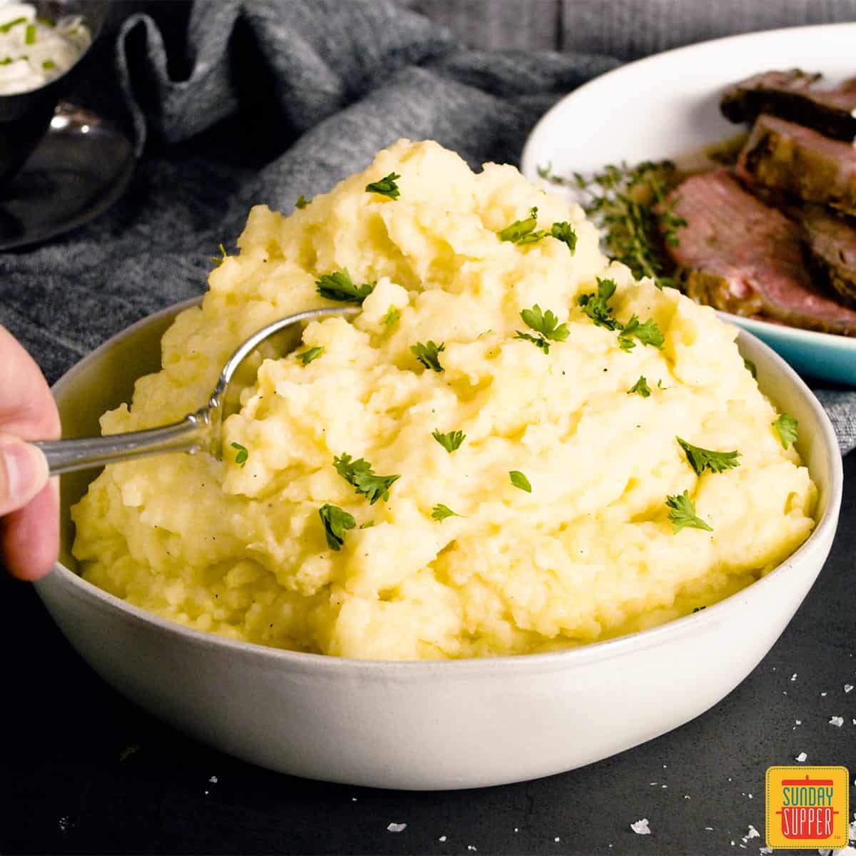 creamy mashed potatoes piled high in a white dish with a spoon and chives