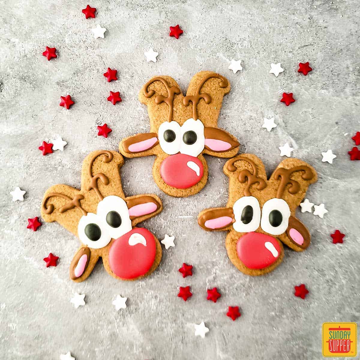 Three reindeer cookies on a surface with star sprinkles