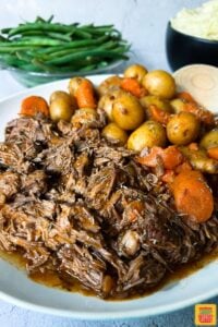 instant pot chuck roast up close on a plate with potatoes and carrots in front of two bowls of green beans and mashed potatoes