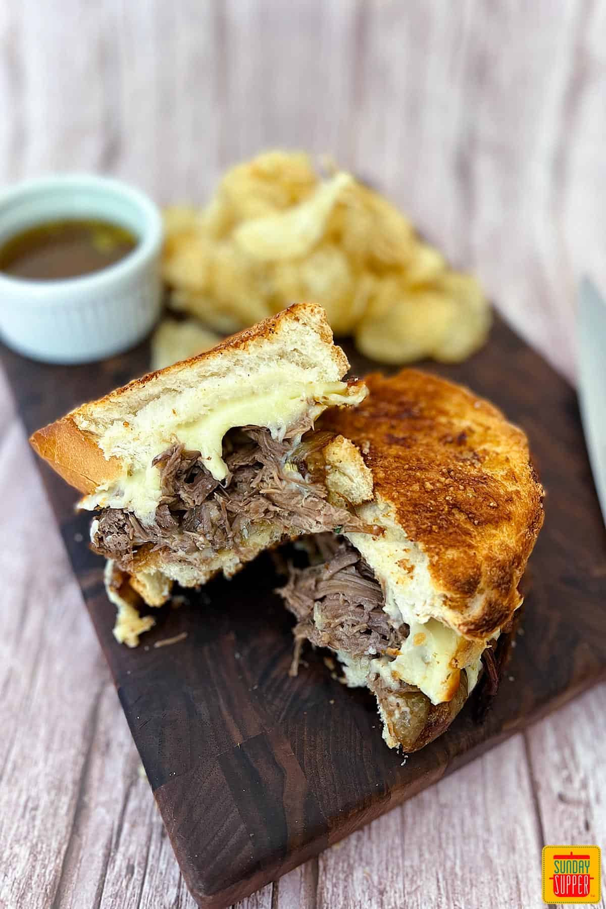 pot roast sandwich sliced in half on a cutting board with chips and au jus