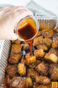 adding bbq sauce to pork belly burnt ends in a disposable pan