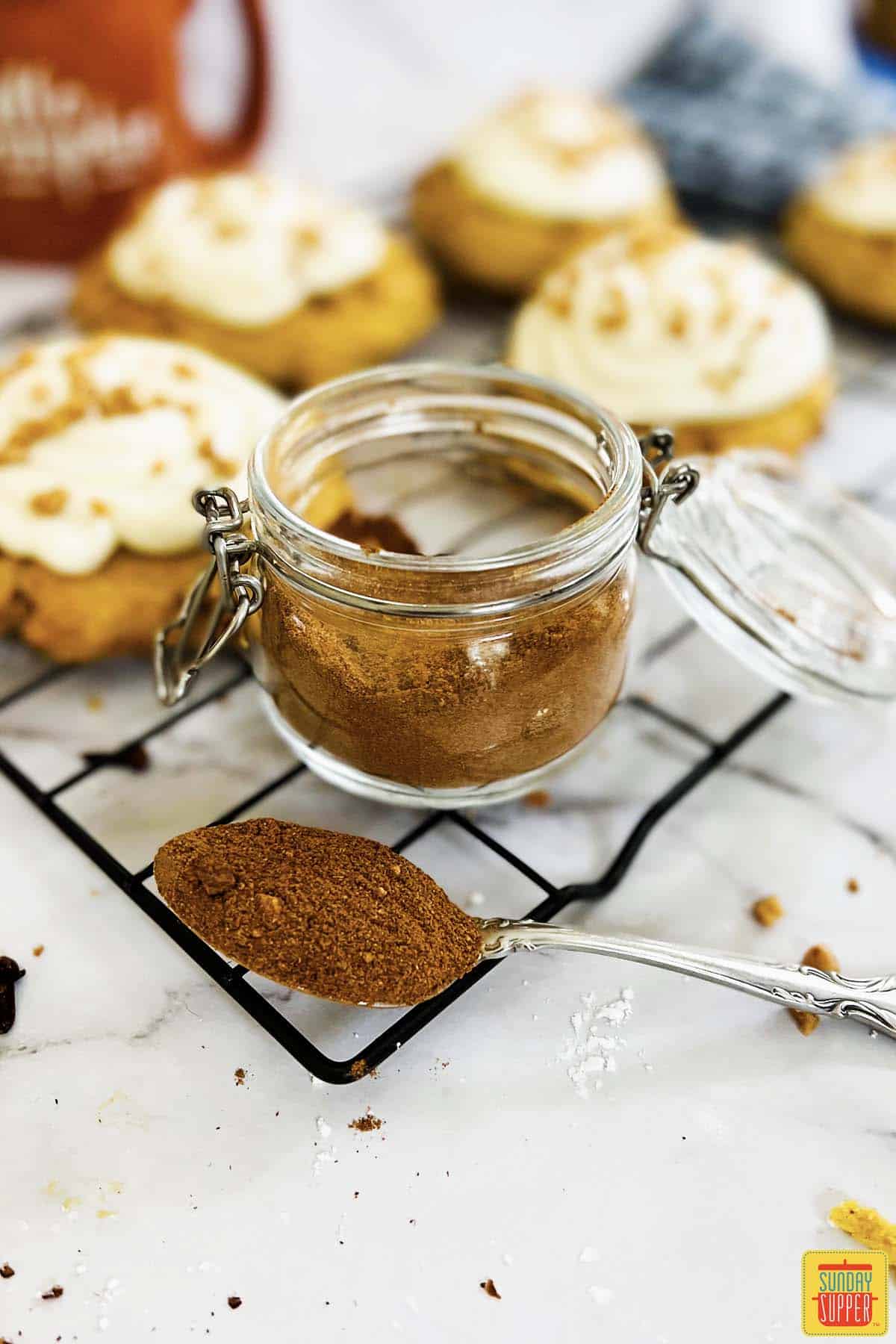 pumpkin pie spice mixed in a jar in front of pumpkin cookies with cream cheese frosting