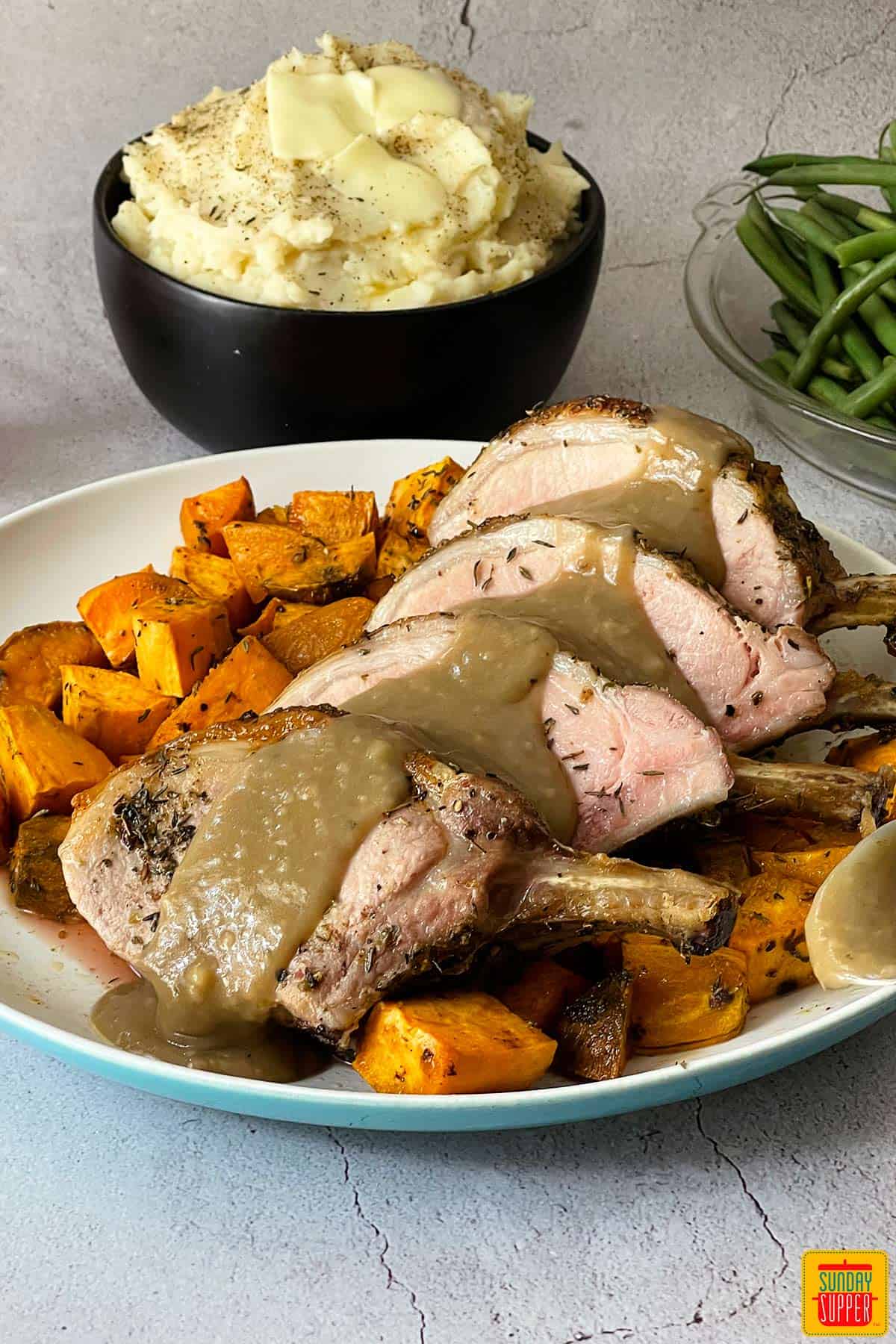 rack of pork sliced with gravy on a white plate with sweet potatoes under neath