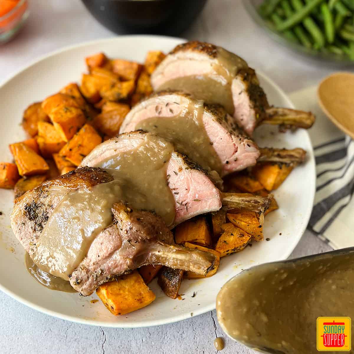 rack of pork sliced on a white plate with gravy over top and sweet potatoes under neath